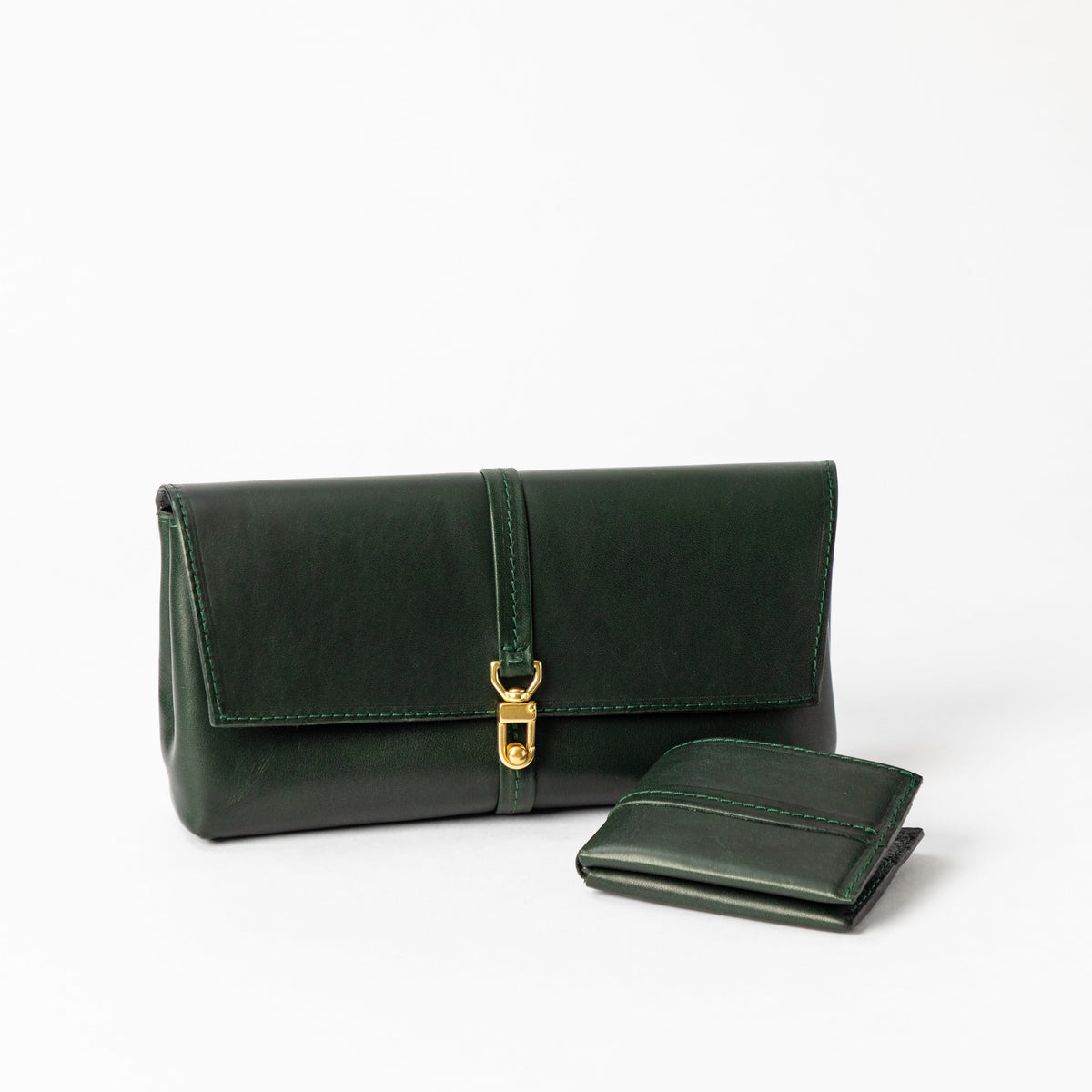 Dark green leather envelope clutch with a wraparound tab and brass clasp beside a matching bifold wallet on white
