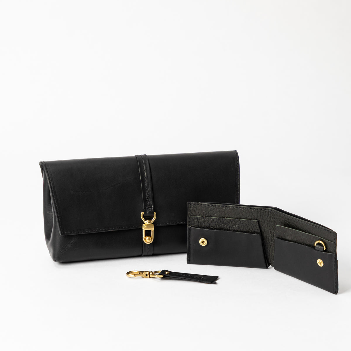 Black full-grain leather bag with brass clasp beside an open bifold wallet and detachable keychain on white