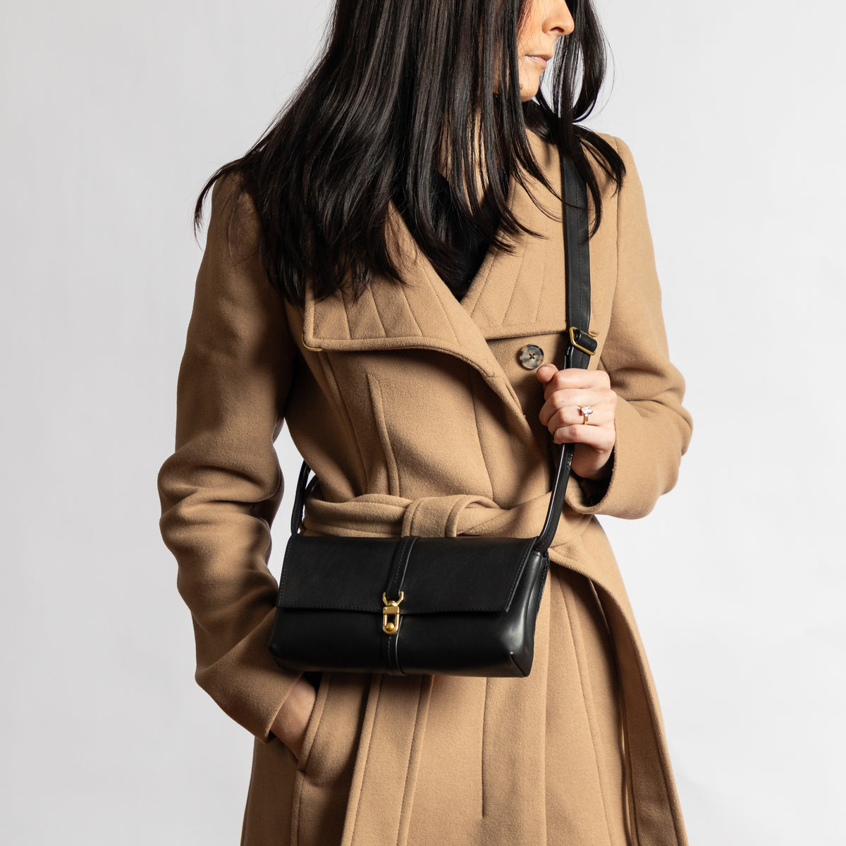 Woman wearing a minimalist black full-grain leather crossbody bag with gold brass hardware and adjustable strap over camel coat