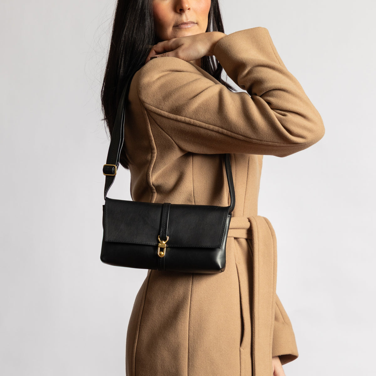 Woman wearing camel wool coat holding black full-grain leather shoulder bag with gold hardware clasp and adjustable strap