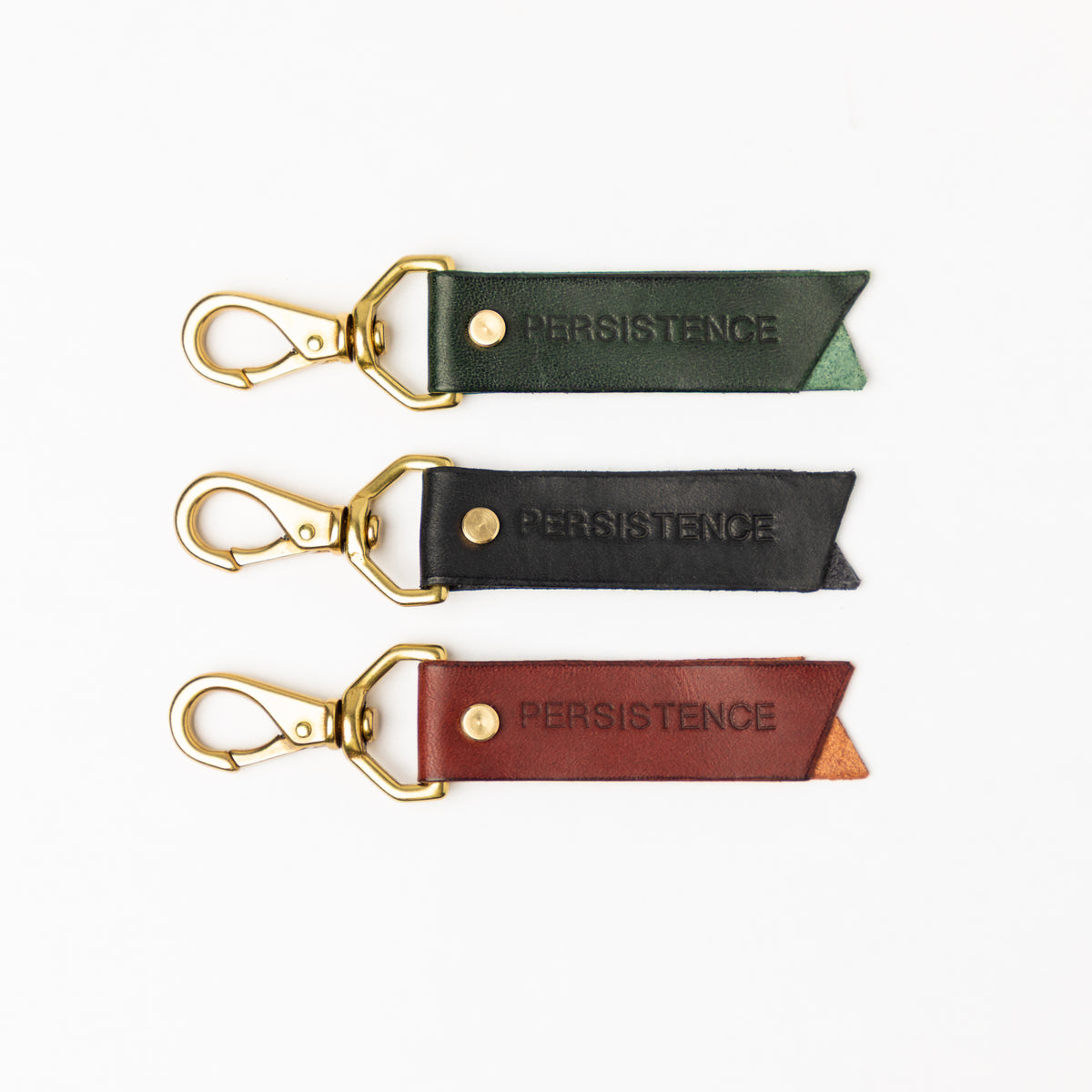 Three PERSISTENCE leather keychains in dark green black and burgundy with brass swivel clasps arranged on white