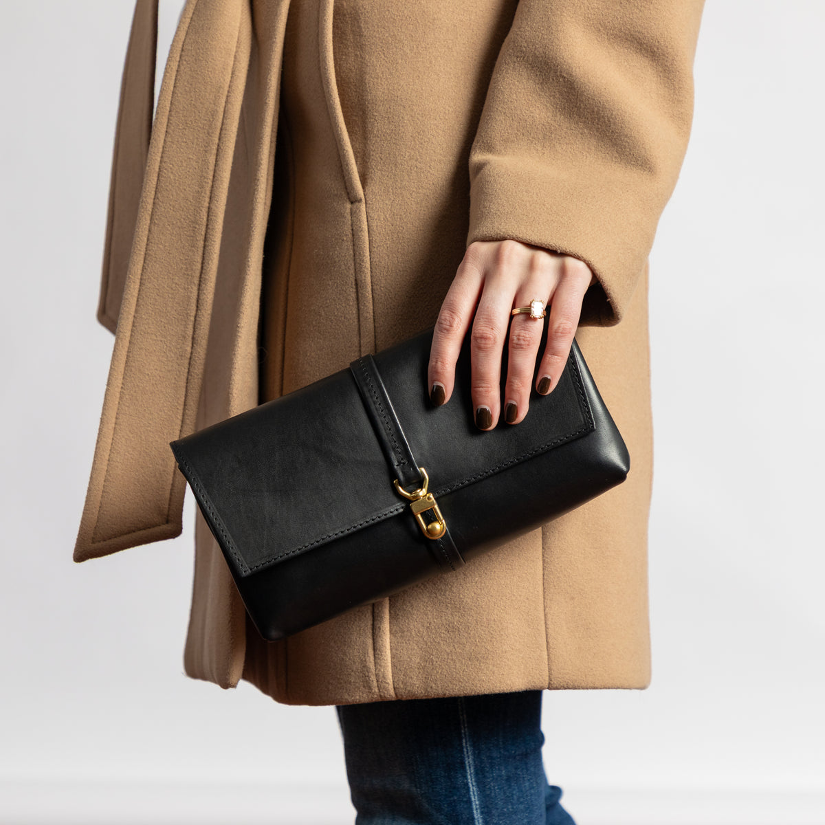Black full-grain vegetable-tanned leather Transpose Bag held as a clutch against a camel coat with polished brass clasp