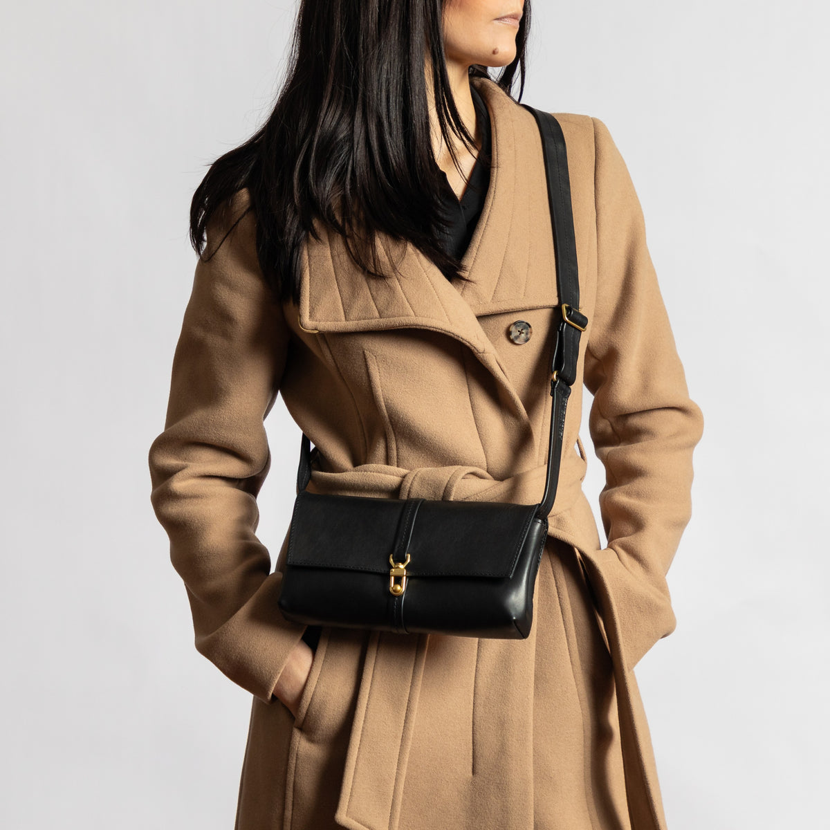 Model wears a black full-grain vegetable-tanned leather Transpose Bag crossbody over a camel coat with solid brass clasp