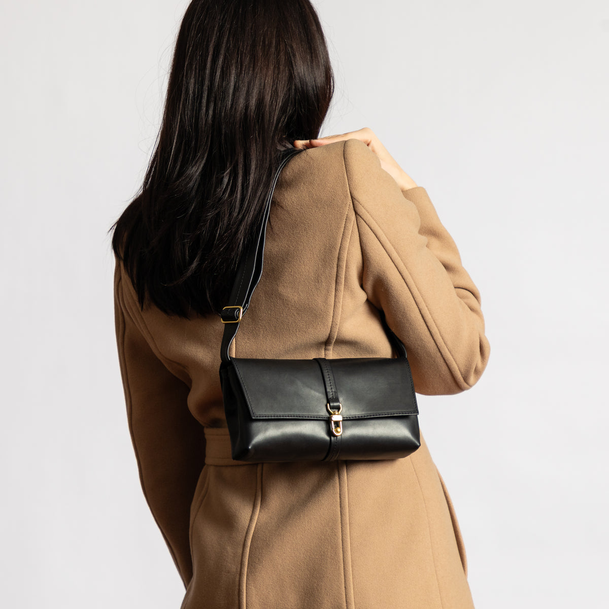 Woman in a camel coat wearing a black vegetable-tanned leather Transpose Bag across her back with adjustable strap and solid brass clasp