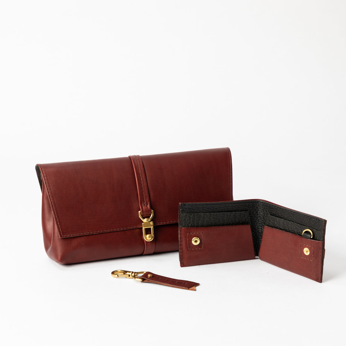 Burgundy leather clutch with brass clasp alongside an open black-trimmed bifold wallet and keychain on white