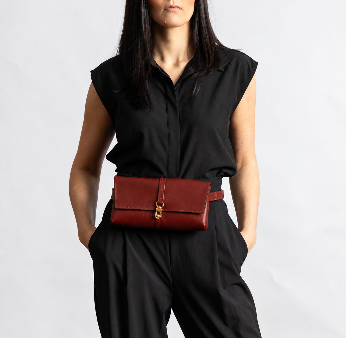 A model wearing a black sleeveless top and black trousers, wearing a dark red (burgundy) leather belt bag with a brass clasp at the waist.