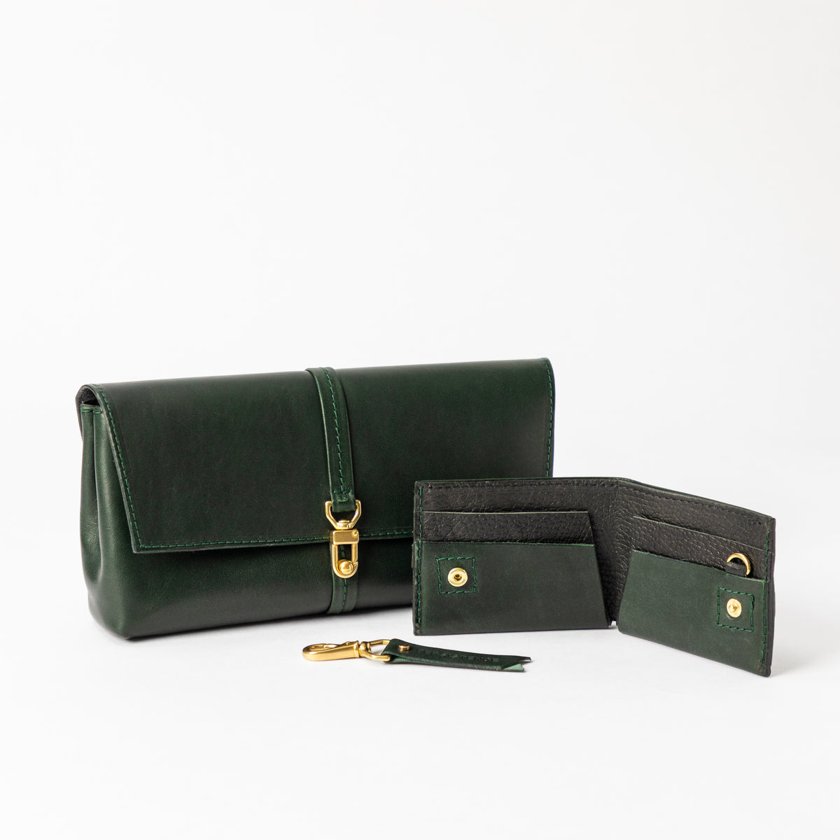 Deep green full-grain leather bag with brass closure behind an open bifold wallet and matching detachable keychain